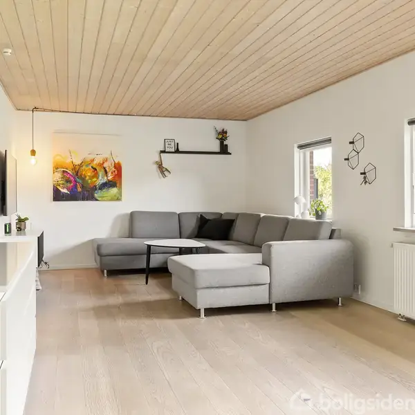 En grå hjørnesofa står i en stue med trægulv og træloft. Vægkunst hænger over sofaen. To vinduer giver naturligt lys. Enkel dekoration og møblering.
