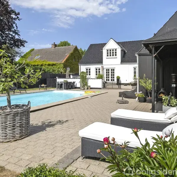 Hvide liggestole står ved siden af en rektangulær pool i en flisebelagt have. Et hvidt hus med sort tag ses i baggrunden omgivet af grønne planter.