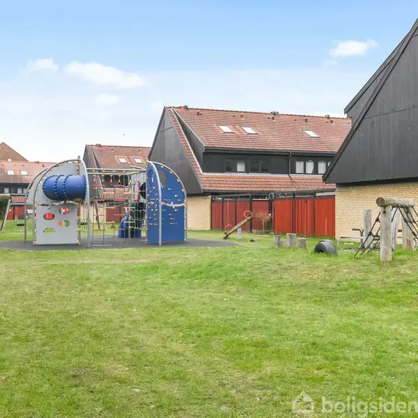 Legeplads med klatrestativ og rutsjebane på en grøn plæne omgivet af rækkehuse med røde tage.