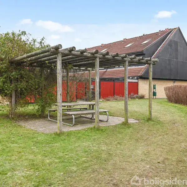 En træpergola med bænk og bord under står på en lille græsplæne. Der er en bygning med rødt tag i baggrunden, omkranset af hække og buske.
