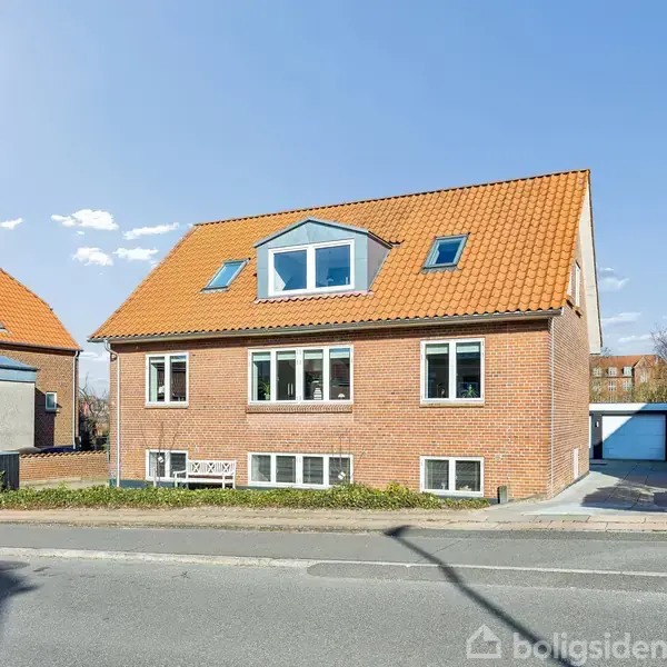 Rødt murstenshus med orangerødt tegltag, centralt placeret på en stille vej. Til venstre og højre ligger nabohuse, og i baggrunden kan trækroner anes.