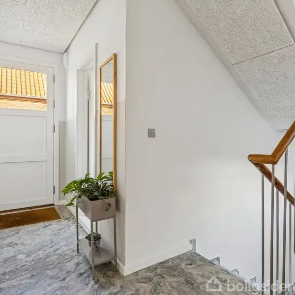 Entré med marmorlignende gulv og trappe opad til højre. Hvid dør for enden, lille plante og spejl langs væggen, der skaber en lys og indbydende atmosfære.