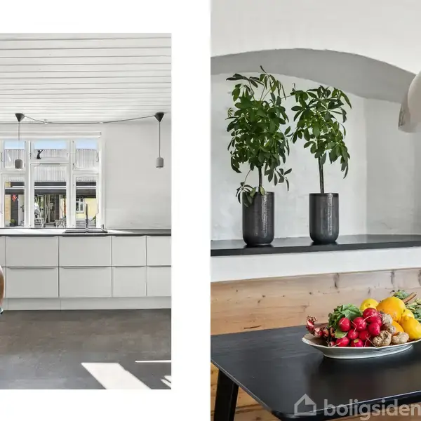 En lys køkkenindretning med hvidt loft og indbyggede skabe, solbelyst bord med planter i sorte potter og en frugtskål som midtpunkt. To vinduer og træstole omkranser rummet.