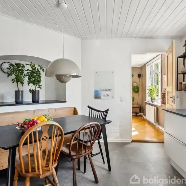 Et spisebord med stole står i et køkken med hvide vægge og loft. En lampe hænger over bordet. Frisk frugt står på bordet, mens planter pryder en hylde i baggrunden.