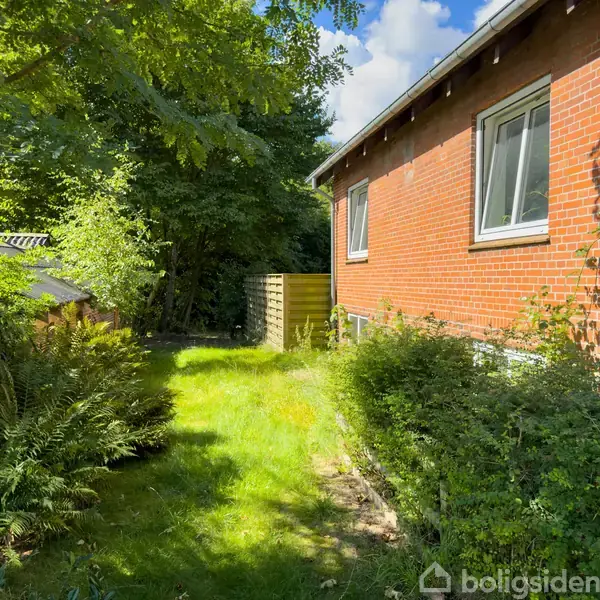 Murstenshus med to vinduer står ved siden af en grøn have, hvor buske og græs vokser tæt op mod husvæggen. Omkranset af træer giver skygge og privatliv.