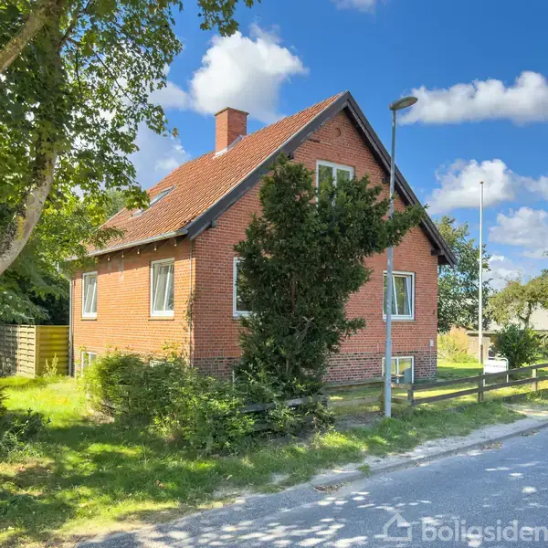 Rødt murstenshus står stille ved en vej omgivet af træer og buske under en blå himmel med skyer. En lygtepæl ses i forgrunden ved husets side.