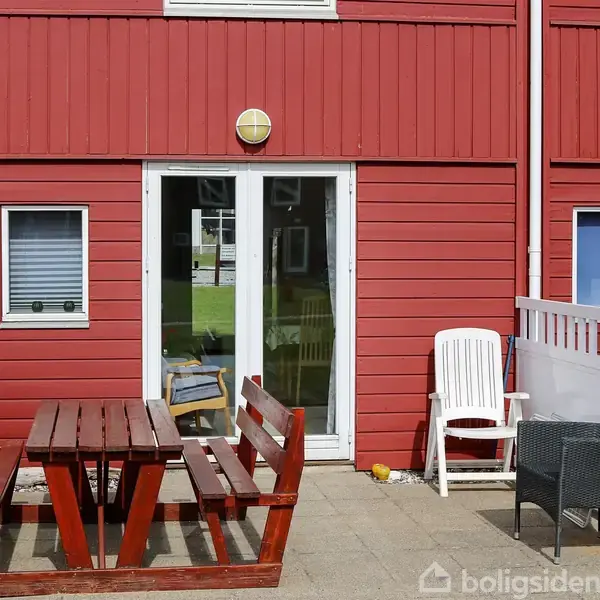Rød træbygning med hvid dør og vindue, omgivet af terrasse med træbord, bænk og plaststole. Terrasse indhegnet med hvide stakit, belagt med fliser.