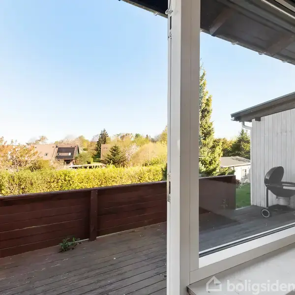 En åben terrassedør viser en træterrasse med en grill i forgrunden. I baggrunden ses græsplæner, træer og et hus under en klar blå himmel.