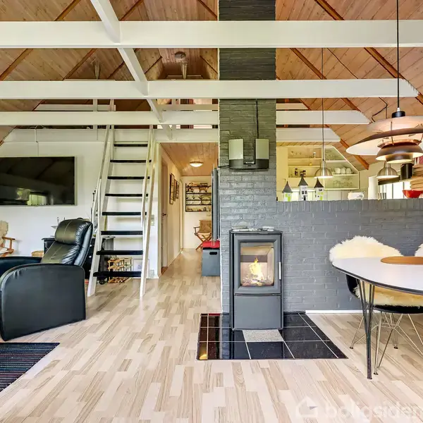 En moderne brændeovn står i et lyst stueområde med loft til kip og trægulv, omgivet af møbler, trapper til hems og et spisebord ved en murstensvæg.