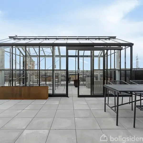 Et glasdrivhus står på en stor, flisebelagt tagterrasse. I drivhuset ses bordplader og stole. Blå himmel og bylandskab er synlige i baggrunden.