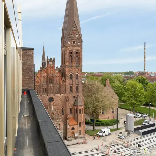 En stor, detaljeret kirke med flere spir står ved siden af en moderne bygning. Den omgivende kontekst inkluderer grønne træer og en klar, blå himmel.
