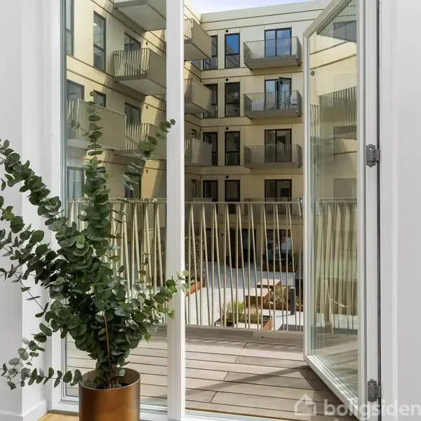 En åben altandør med udsigt til en gårdhave omkranset af boligkompleksets facader. En grøn plante står ved døren, og altanen har en metalrækværk.