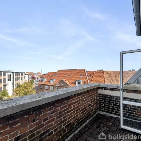 Åben altandør i murstensvæg åbner op til en altan med udsigt over rødt tegltag og moderne bygninger i baggrunden under en blå himmel.