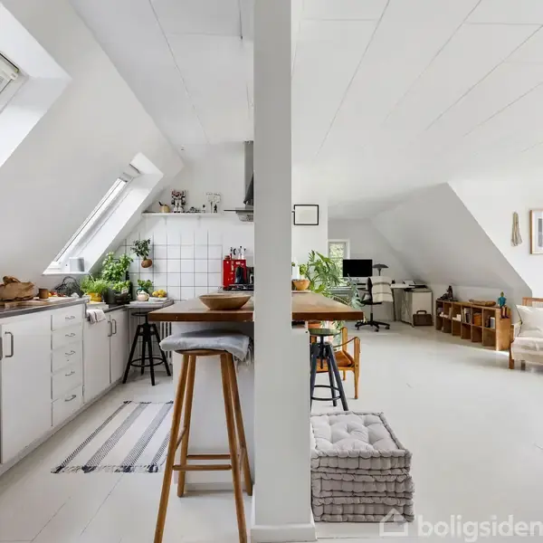 Et lyst køkken-alrum med skråvinduer og hvide skabe. Der står en barstol ved bordet, og flere planter pynter rummet. I baggrunden ses en stue med sofa og billeder.