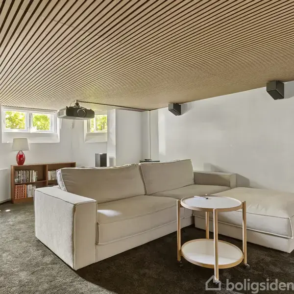 En hvid sofa står i en moderne stue, omgivet af et lavt træloft og små vinduer. Der er et lille bord ved sofaen, og bagerst står en reol.