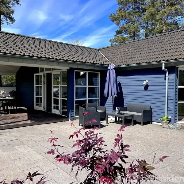 Et træhus med blå facade har en terrasse med havemøbler og en parasol. Omgivet af træer under en blå himmel.