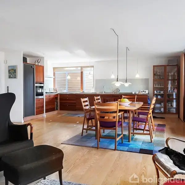 Stue med spisebord og seks træstole står på farvet tæppe. Til venstre en sort lænestol med fodskammel. Store vinduer giver masser af lys. Appendix kitchens vises i baggrunden.