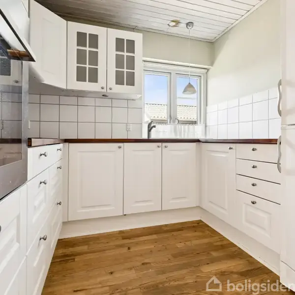 Hvidt køkken med lyse trægulve. Skabene har sølvfarvede håndtag. Et stort vindue med udsigt giver naturligt lys. Over vasken hænger en enkel lampe. Tiles pryder væggene.