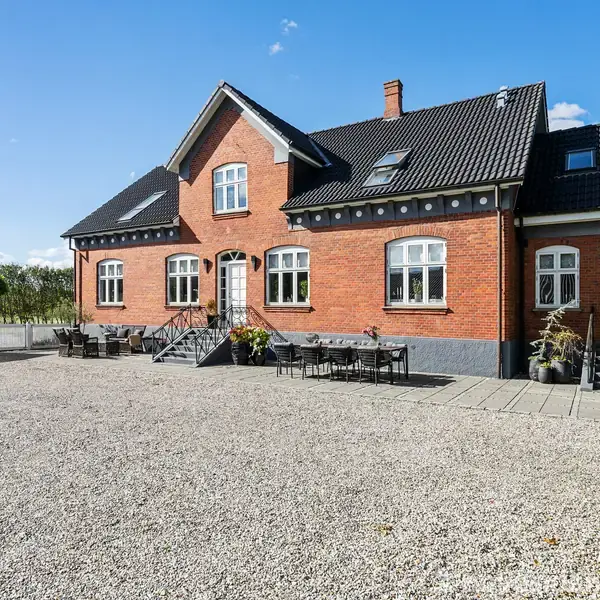 Rødt murstenshus står på grusplads, omgivet af buske og grønne marker. Huset har sorte tagsten, flere vinduer og trapper, og foran er placeret havemøbler.