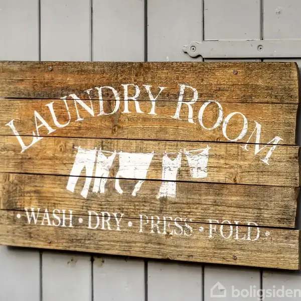 Træskilt med teksten "LAUNDRY ROOM" og "WASH - DRY - PRESS - FOLD" hænger på en grå væg. Skiltet prydes af grafik af tøj på en tørresnor.