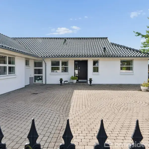 Et hvidt hus med sort tegltag står midt i et brolagt område. Flere vinduer ses på facaden, og planter flanker indgangen. Blå himmel og træ ses i baggrunden.