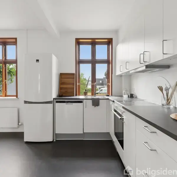 Et moderne køkken ligger til venstre med hvide skabe og komfur, ved siden af en plante i potte. Til højre er en stue med sofa, stole og trægulv.