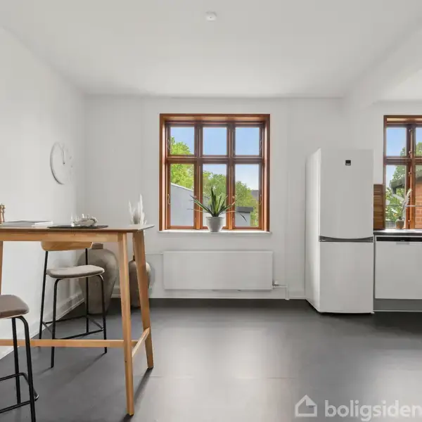 Et højt spisebord med to stole står i en lys, moderne køkkenalrum. Køleskab og køkkenskabe er til højre. Store vinduer i baggrunden giver udsigt til grønne træer.