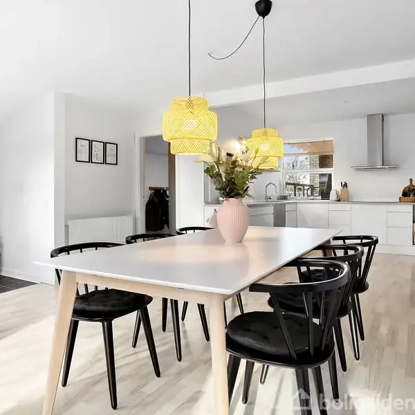 Spisebord med sorte stole står i et lyst køkken-alrum. Over bordet hænger to gule pendellamper. I baggrunden er en kamin ved siden af en grå stenvæg.