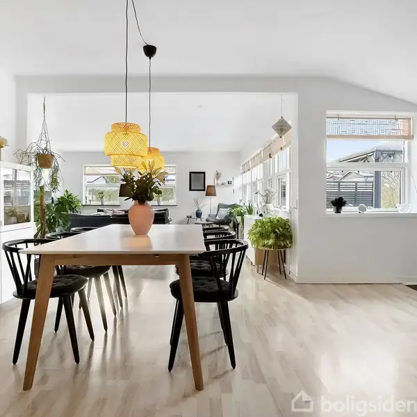 Et moderne spisestuebord med seks sorte stole står på et lyst trægulv. Over bordet hænger to lamper. I baggrunden er der flere planter og store vinduer med naturligt lysindfald.