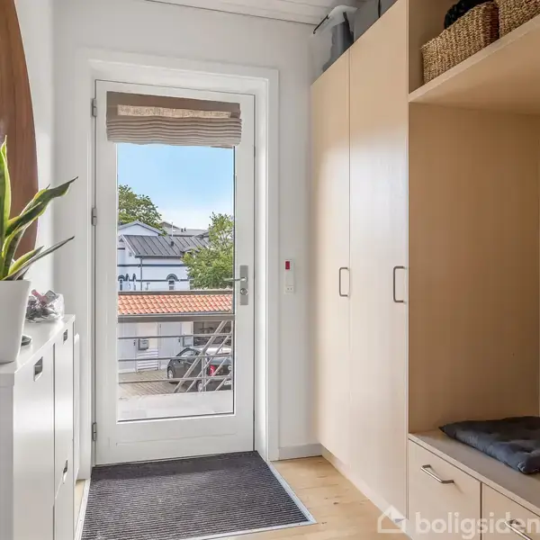 En lys entre med træskab og bænk på højre side, en plante på hvid kommode til venstre, og en glasdør i baggrunden viser et hus med tegltag udenfor.