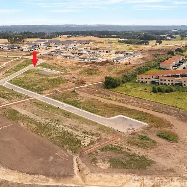 Grønt område med byggegrunde, markeret med rød pil. Nye boligbyggerier ses i baggrunden. Landligt område med marker og spredte træer omkring.