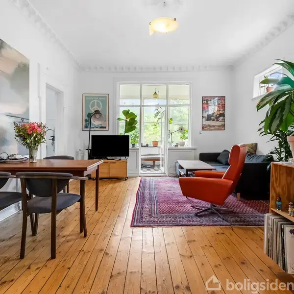 En stue med trægulv indeholder et spisebord med blomster, en rød stol, sofa, store planter og en reol med plader. Væggene har kunst, og der er et vindue i baggrunden.