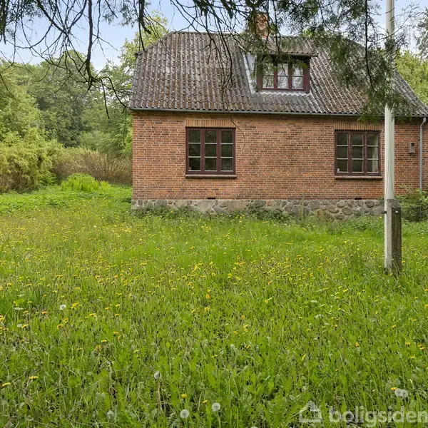 Et rødt murstenshus står stille i en grøn have med højt græs og buske. Baggrunden domineres af træer, og et andet hus skimtes til venstre.