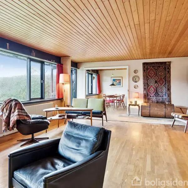 En stue indrettet med sorte og grønne lænestole, en bogreol og glasvægge. Rummet har trægulv og træloft samt udsigt til en spisestue. Røde gardiner og lamper skaber varme.