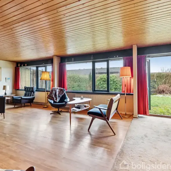 Stue med trægulv og træloft indeholder moderne møbler og store vinduer med udsigt til en have. Rummets lyse indretning inkluderer lamper og røde gardiner, der skaber en varm atmosfære.