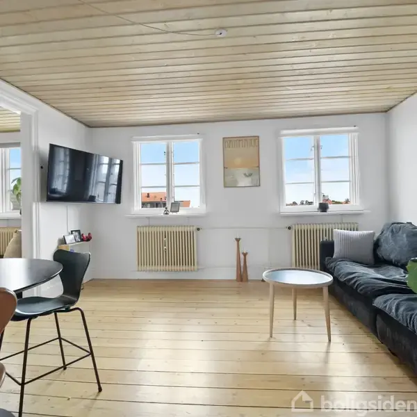 Stue med sort sofa langs en hvid væg, to vinduer med udsigt, og et væghængt TV. Rundt sofabord står på trægulv. I forgrunden ses et spisebord og stole.