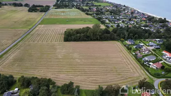 En stor mark med striber fra nyligt høstet afgrøde ligger ved siden af en landsby med huse; skovbeklædte områder ses i baggrunden nær kystlinjen.