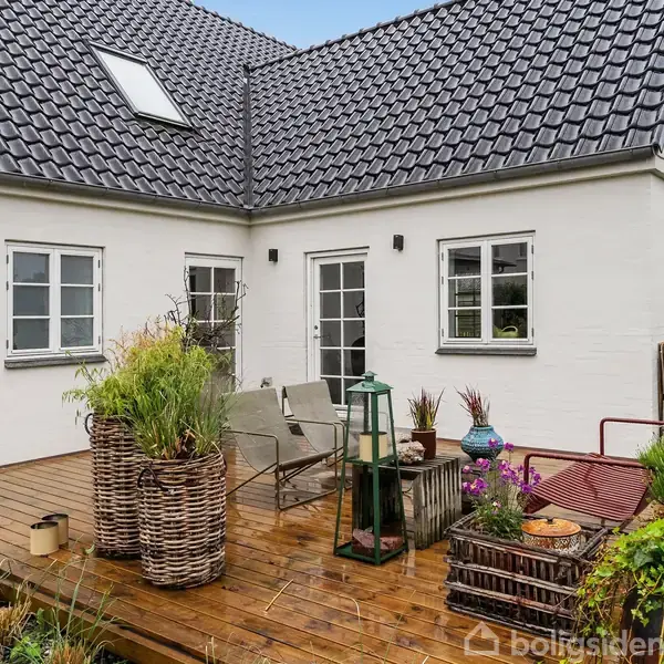 Et hus med hvid facade og mørkt tag står med en træterrasse foran. På terrassen er der kurvemøbler, planter og en lanterne, omgivet af en have.
