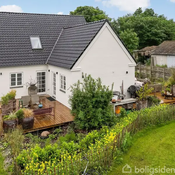 Et hvidt hus med mørkt tag står på en frodig grund. En træterrasse foran er udsmykket med planter og havestole. Til venstre ses et skilt med teksten "SAMSØ".