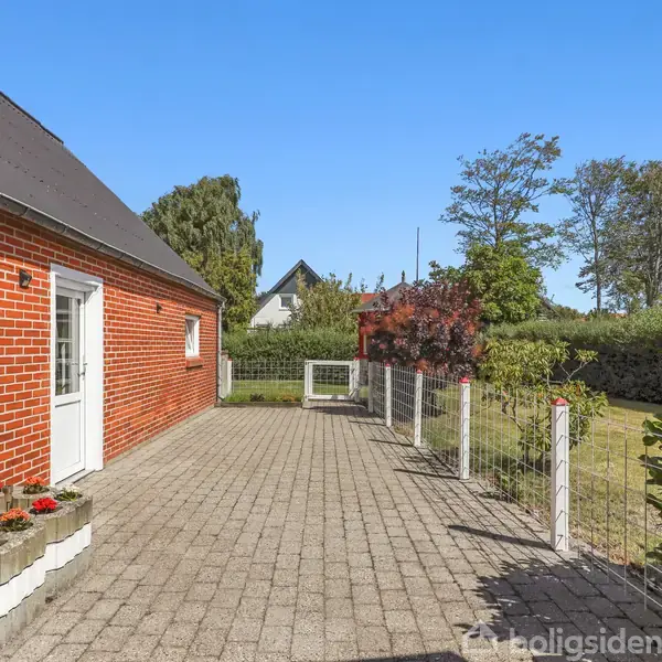 Røde murstenshus med hvide vinduer og dør. Flisebelagt terrasse foran huset omkranset af lavt hegn. Haven er frodig med buske og træer, under blå himmel.