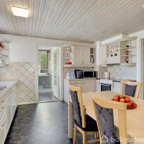 Køkken med hvidt loft og skabe, spisebord med stabel af frugter. Flisevægge, mørkt gulv, mikrobølgeovn og ovn. Dør åbner til andet rum med vindue mod grøn udsigt.