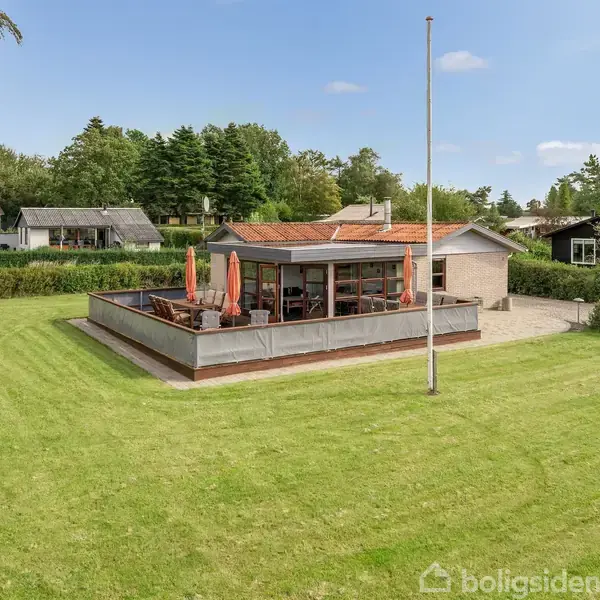 Et enfamiliehus med en stor terrasse ses i en grøn have med velplejet græsplæne og flagstang. Terrasse har parasoller, omgivet af træer og nabobygninger i baggrunden.