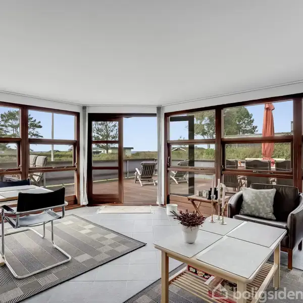 Stue med sofa og sofabord i forgrunden, store vinduer mod en terrasse med udsigt til træer og en have. Rundt spisebord med stole står ved siden af.