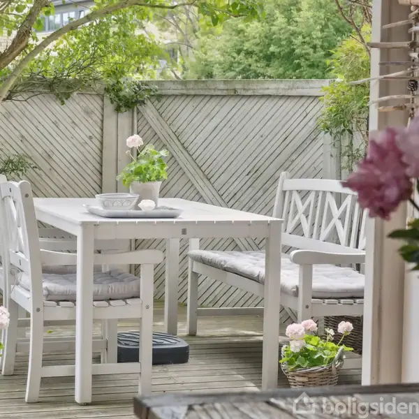 Hvid havebord med stole er opstillet på en træterrasse omgivet af grønne planter og et hegn. Pottede blomster dekorerer området, mens lyserøde blomster ses i forgrunden.