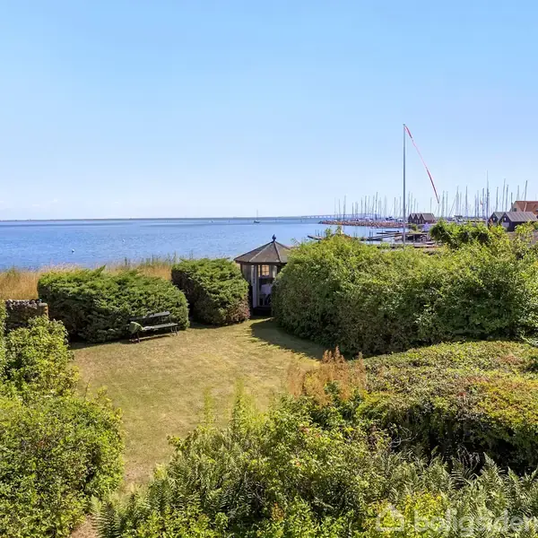 Pavillon står stille midt i en frodig have omgivet af tætte buske. I baggrunden ses en rolig havudsigt med både fortøjet ved en havn.