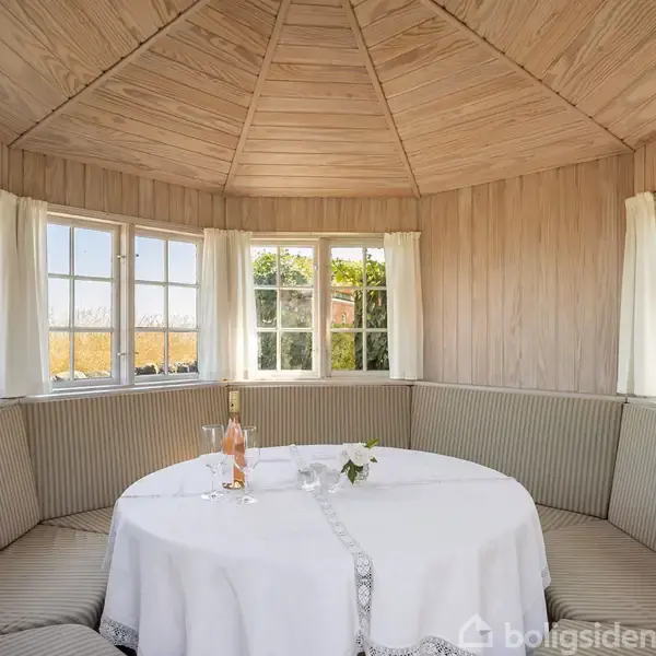 Rundt bord dækket med dug står i midten af en lys pavillon med trævægge og polstrede bænke. Flere vinduer omkranser rummet, hvilket giver udsigt til grønne omgivelser.