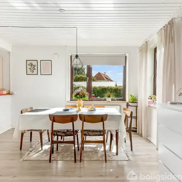 Et spisebord med dug og fire stole er placeret centralt i en lys stue med stort vindue. Bordet har tallerkener og frugt. Indretningen er moderne og indbydende.