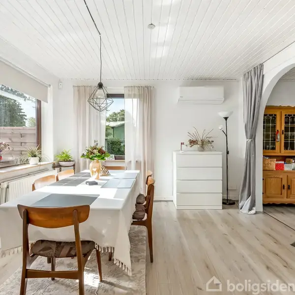 Spisebord med fire stole dækket med en hvid duge står under en moderne lampe i et lyst rum med hvide vægge og træloft. Skænk, planter, store vinduer og kommode ses.