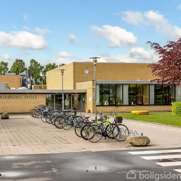 Skolebygning med skiltet "SAKSKØBING SKOLE," omgivet af parkerede cykler på en flisebelagt plads. Flere træer og en græsplæne ses i baggrunden under en blå himmel med skyer.
