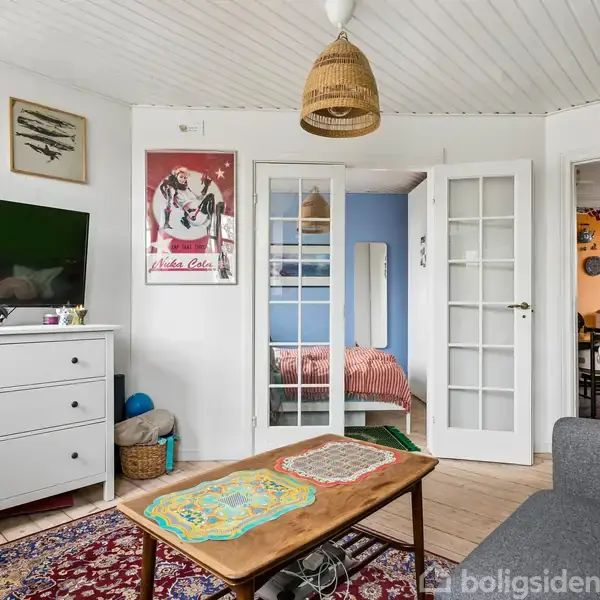 Stue med grå sofa og træbord, dekoreret med farverige tæpper. Fjernsyn står på en hvid kommode. To franske døre fører til soveværelse. Vægkunst inkluderer en plakat og flere billeder.